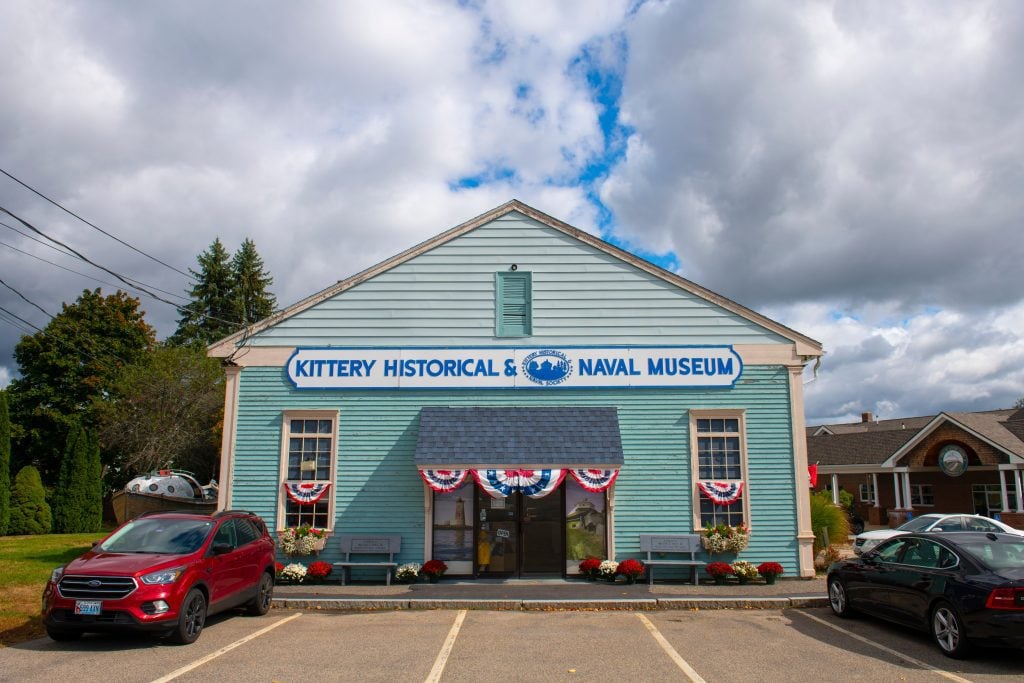 A small blue cottage with a sign reading Kittery Historical & Navy Museum, with patriotic red white and blue paper banners hanging above the door.