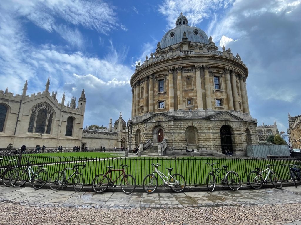 A round tower at Oxford University, on a green surrounded by a fence with lots of bikes locked to it.