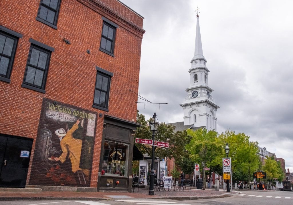 Downtown Portsmouth New Hampshire, with a small town square featuring a tall white steepled church, and a brick building with a mural of Italy on it.