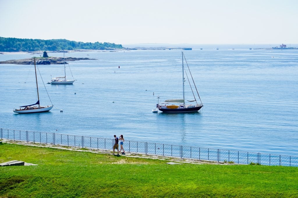 A park on the edge of the river, several sailboats in the water behind it.