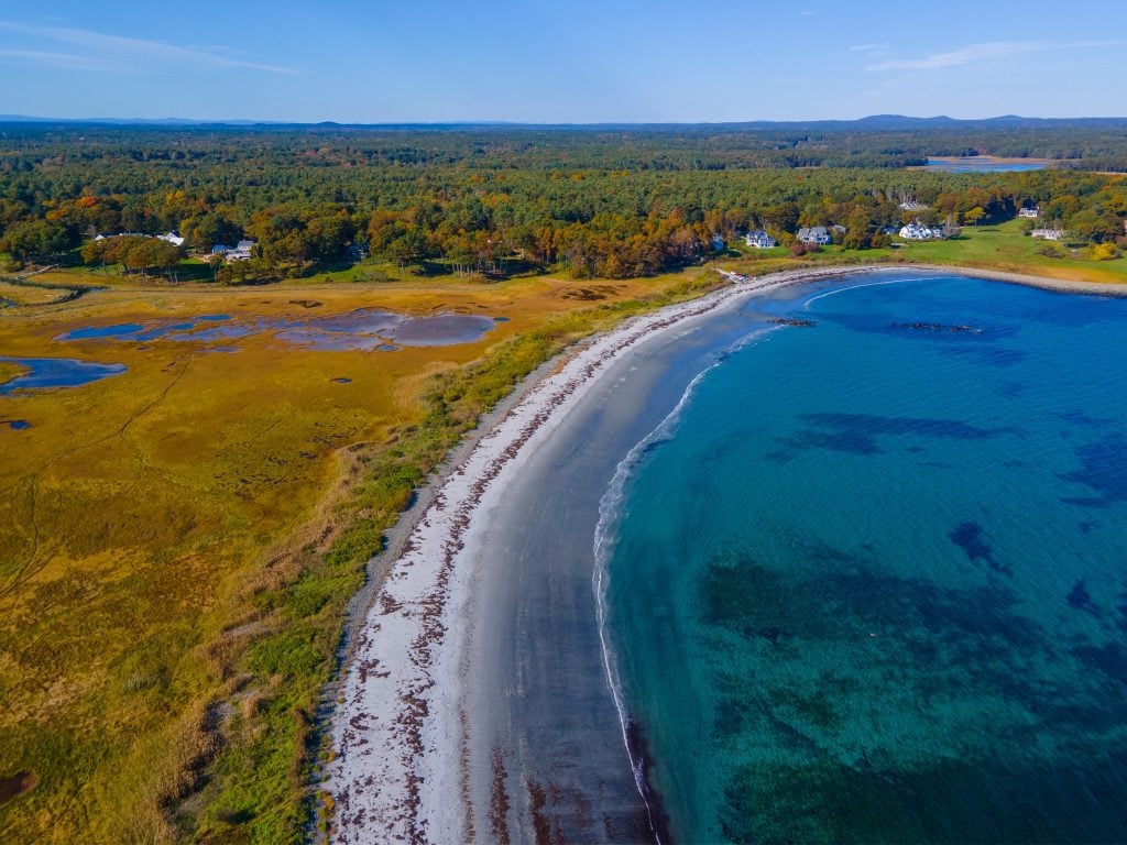 An Aerial view of a gray sand beach curving around bright blue water, with wetlands on the other side.