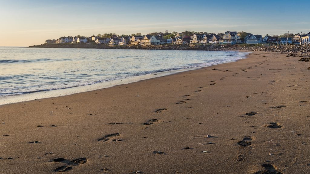 A calm beach at sunrise in Maine. In the background, a row of cottages (that look modest but are probably multi-million dollar homes) along the coastline.