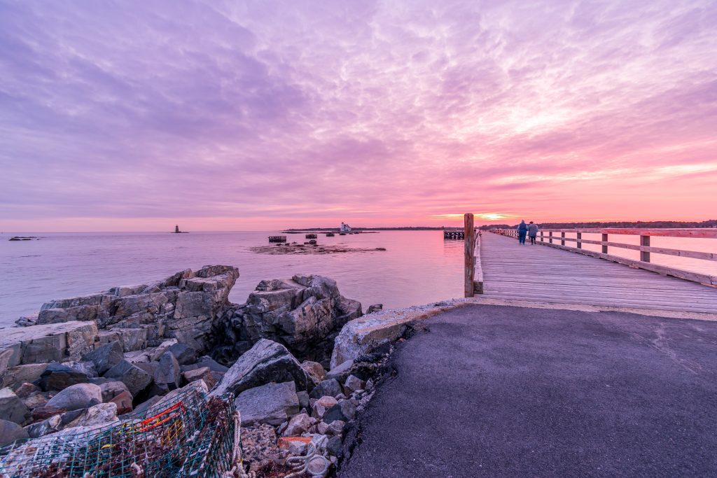 A wooden pier leading into the water in Maine, underneath a pink and purple sunset.