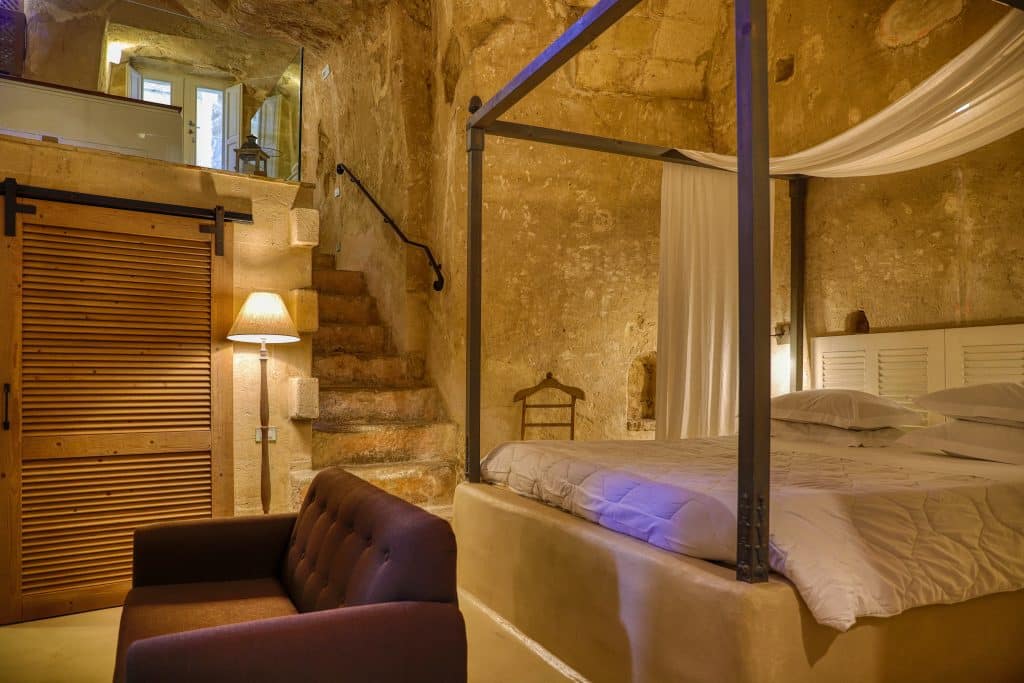 A luxury hotel room inside a cave, with a four-poster bed and couch, all surrounded by sand-colored stone walls.