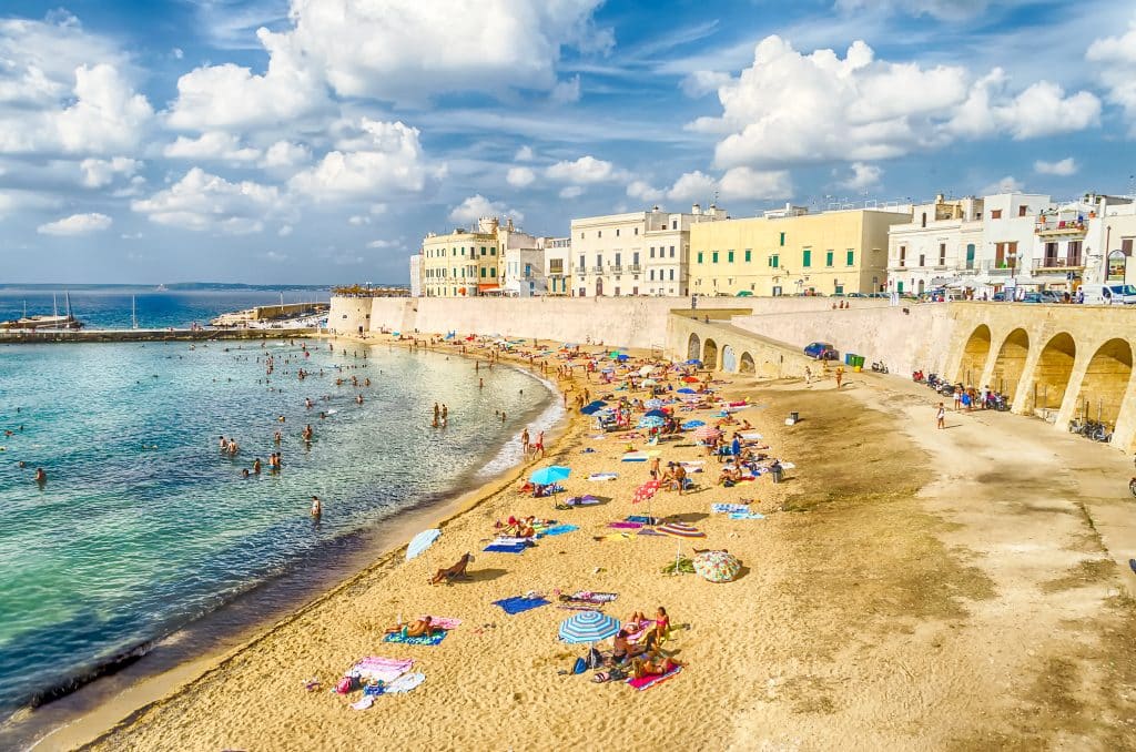Gallipoli, Italy, a city of pastel buildings perched on a soft sand beach with people sunbathing on it, next to clear turquoise water.