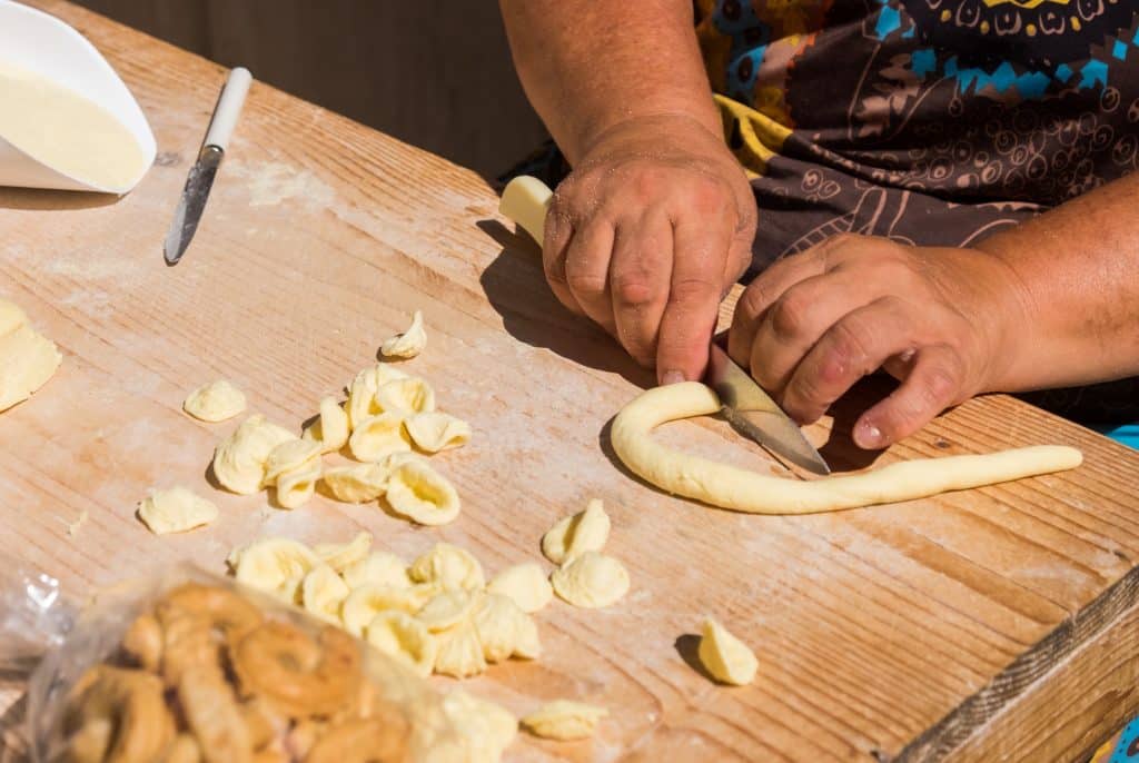 A woman's hands rolling out a long skinny tube of pasta dough with her knife ready to cut it into ear-shaped pieces.