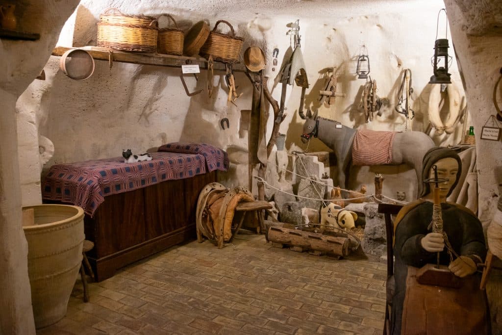 À l'intérieur d'une maison troglodyte à Matera : un petit lit dans un coin, de nombreux outils accrochés aux murs, un âne empaillé et un mannequin représentant une femme en train de filer de la laine.