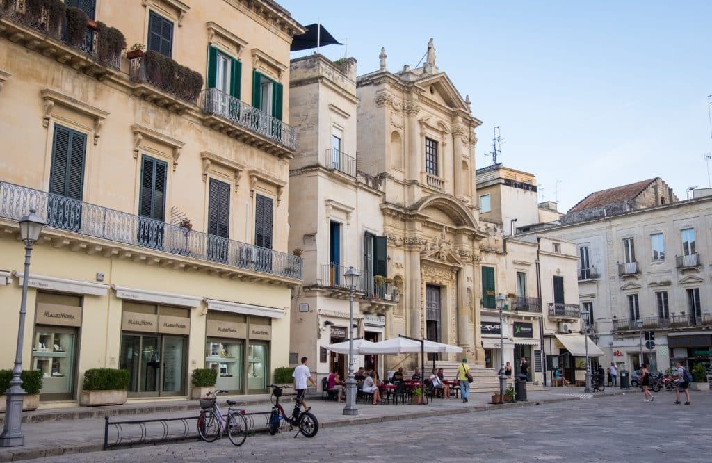 A street in Lecce with a mix of baroque and modern buildings, street cafes below, bicycles parked nearby.