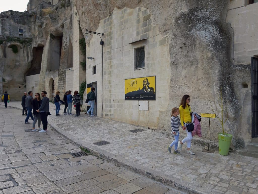 People walking down a stone street in Matera next to a church with a Dali exhibit.