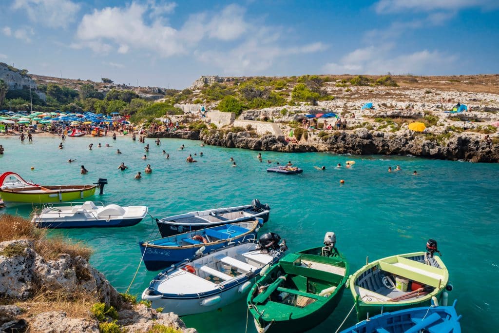 A city perched on bright turquoise sea, wooden rowboats and people swimming in the water.