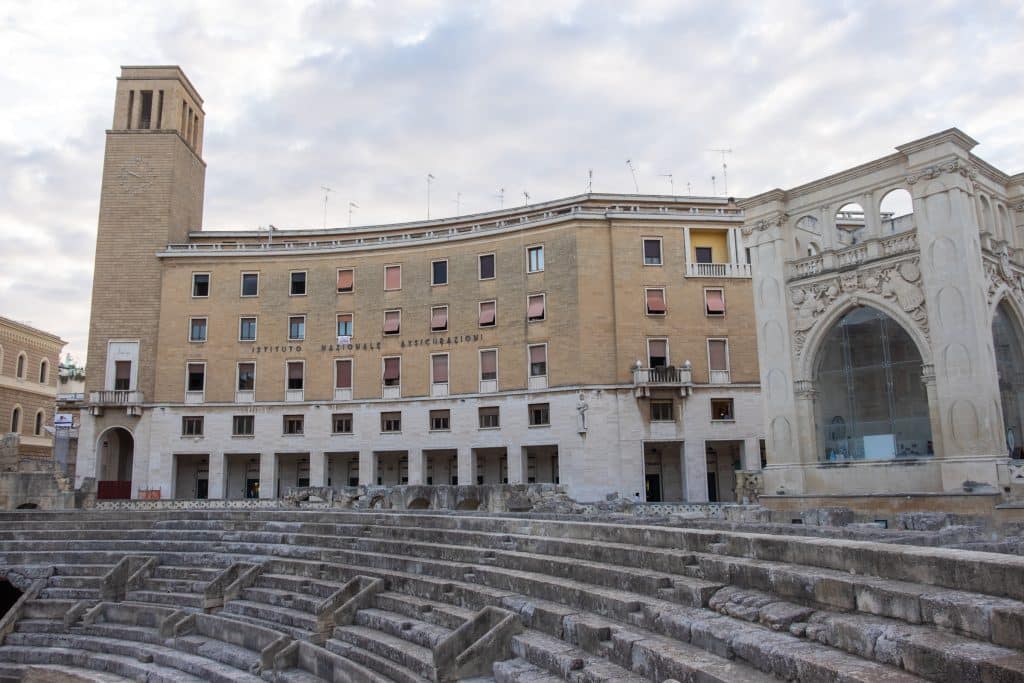 The rounded steps of an ancient Roman amphitheater in Lecce, in front of a fascism-era modern stone building with a clock tower with a modern face on it.