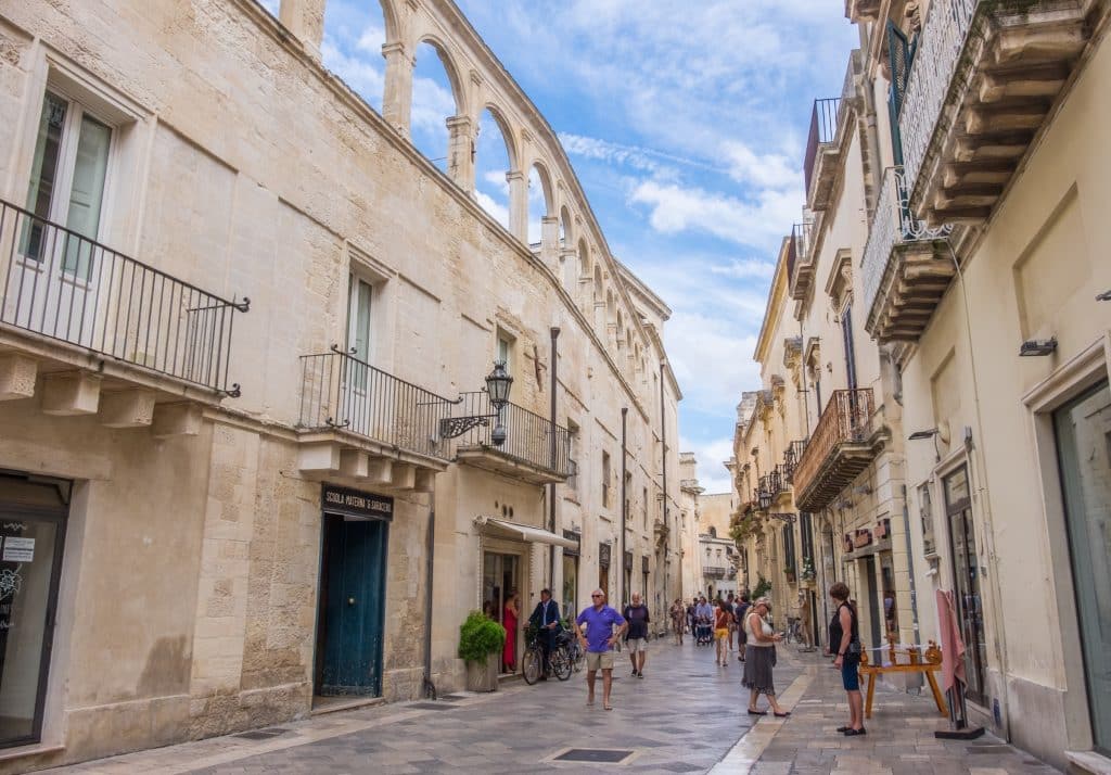 A crowded street in Lecce, Italy, with stone buildings topped  with wrought-iron balconies.