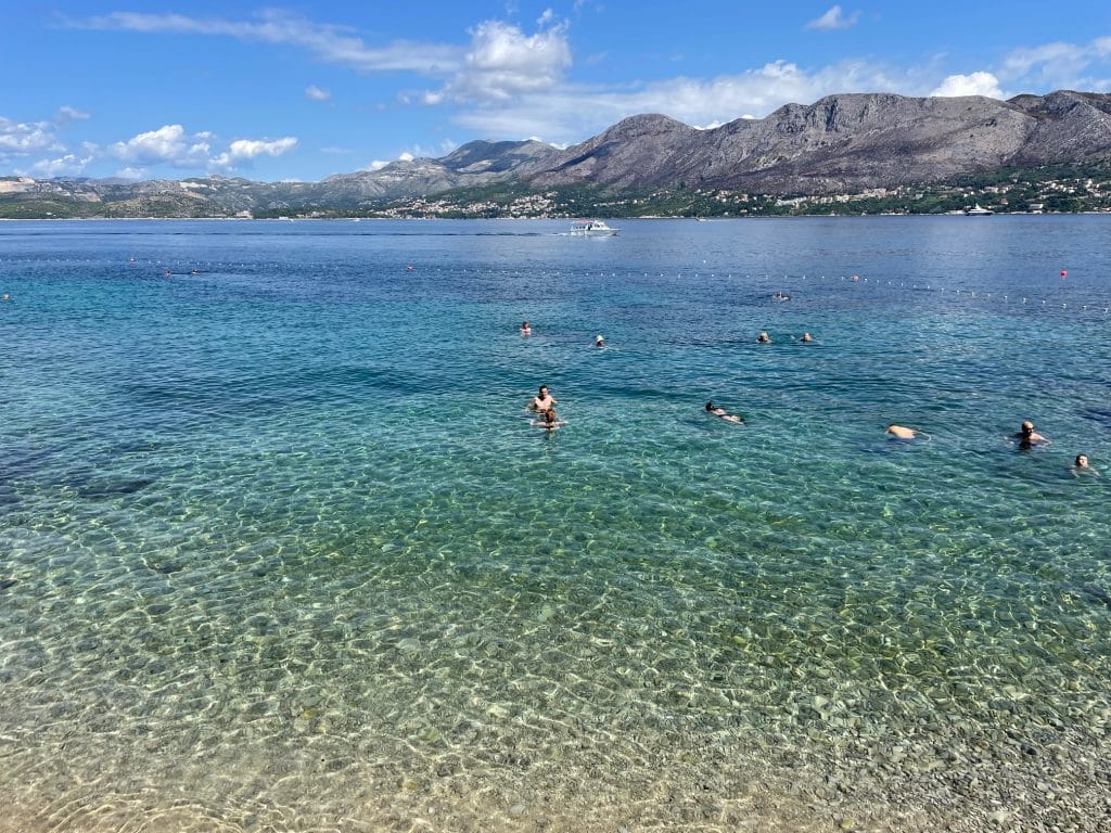 People swimming in the bright green, clear Adriatic Sea, mountains in the background.
