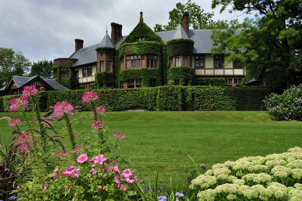 A gilded age mansion with lots of ivy-covered brick walls and turrets, hidden behind shrubbery.