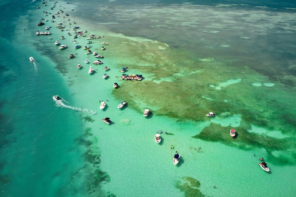 Dozens of small boats anchored off a sandbar in clear, bright green water.