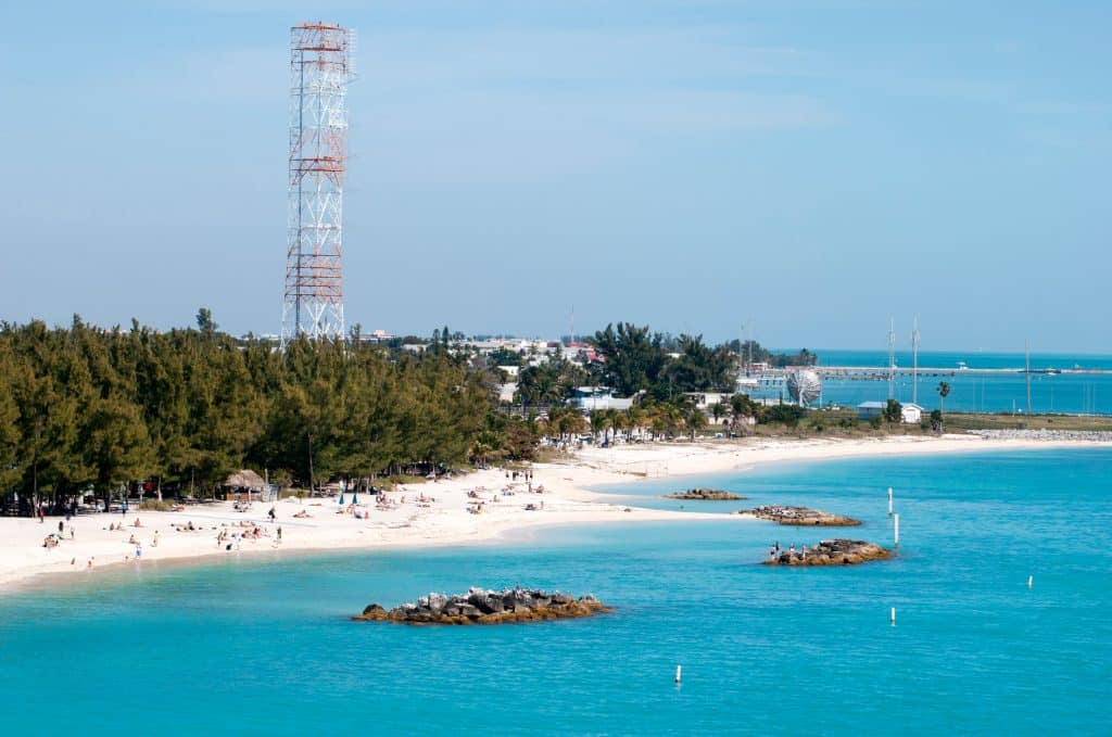 A long white sand beach in front of the blue sea, tons of people sitting on the beach. There is a forest and an signal tower just past the beach.