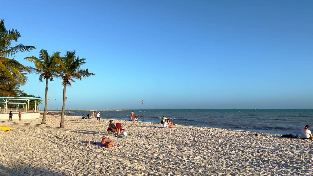 A calm sandy beach at sunset, people soaking up the sun with long shadows extending behind them.