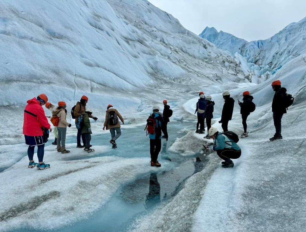 A group of people in helmets walking on a huge blue-white glacier with liquid puddles on it.