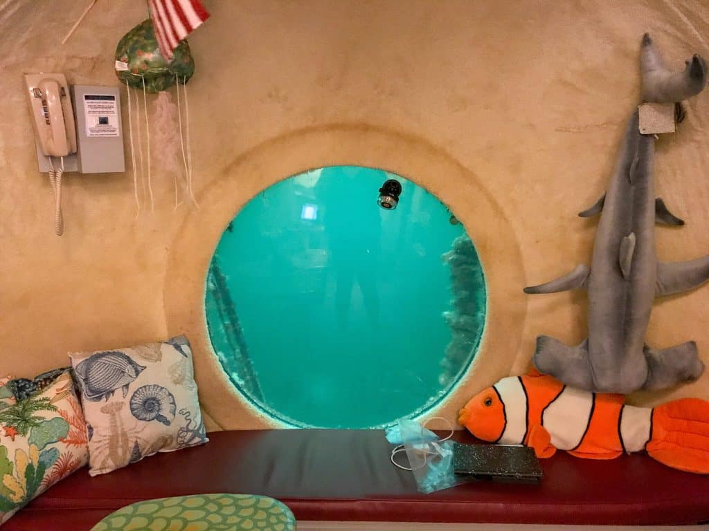 A room in an underwater hotel with a porthole looking into the water, and a bench in front with a stuffed blowfish and some pillows.