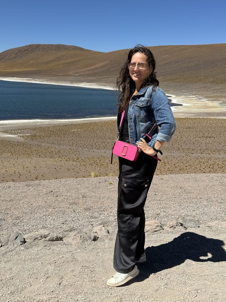 Kate standing in front of a calm lake in the desert, wearing black satin cargo pants, a denim jacket, and a small hot pink pill-shaped purse hanging on her shoulder.