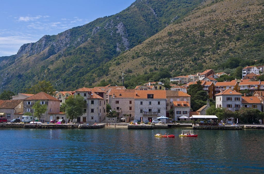 Two people kayaking in the calm Bay of Kotor, white stone buildings with orange roofs on the shore behind them, and mountains rising up in the distance.