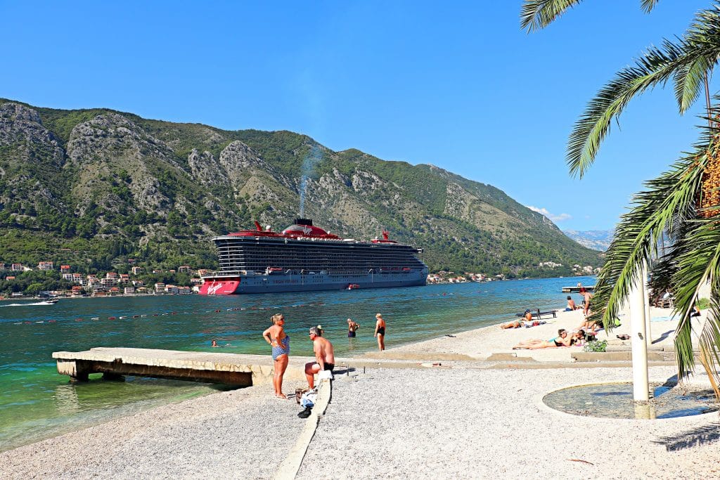 A small, rocky beach on the edge of the calm, Green Bay of Kotor in Montenegro, a giant cruise ship passing by.