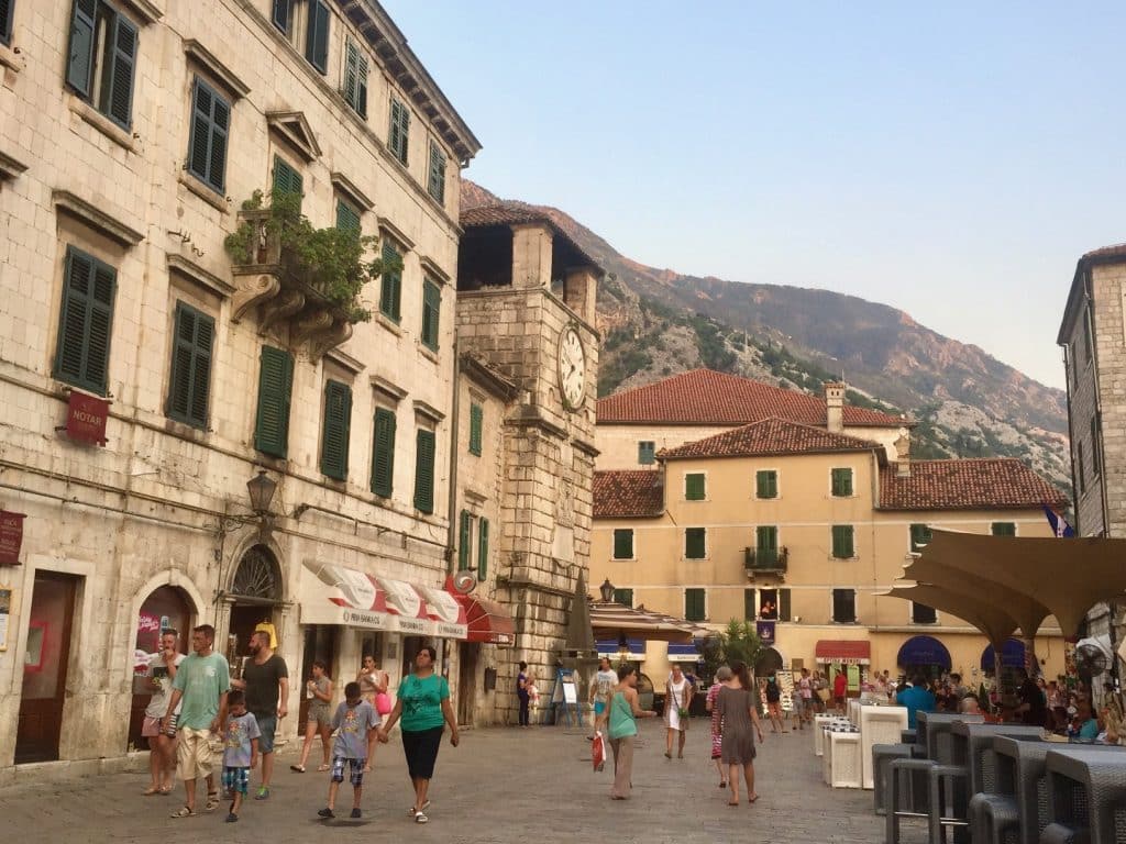 People walking through an old town of stone buildings with green shutters, including a big square clock tower.