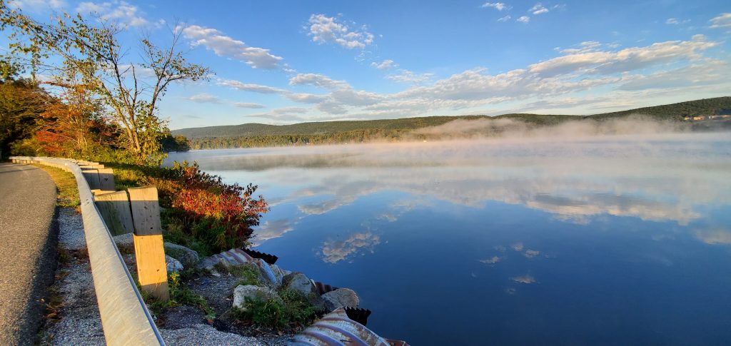 A lake covered with gentle mist on an early fall morning, a partly cloudy sky ahead.