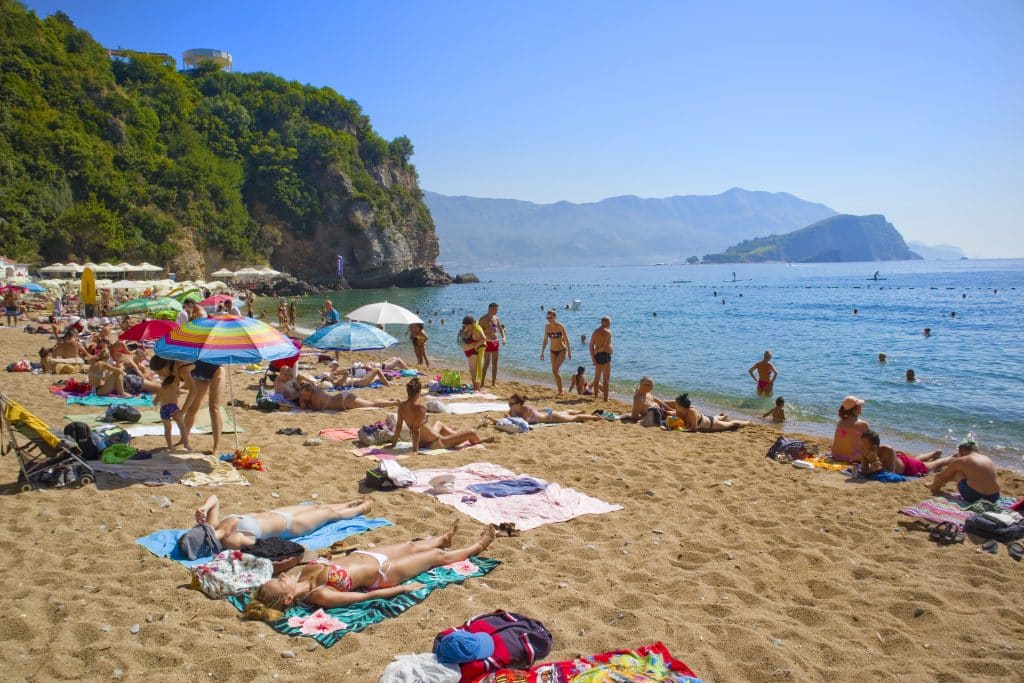 Lots of people lounging on towels on a beach in Montenegro, mountains in the distance.