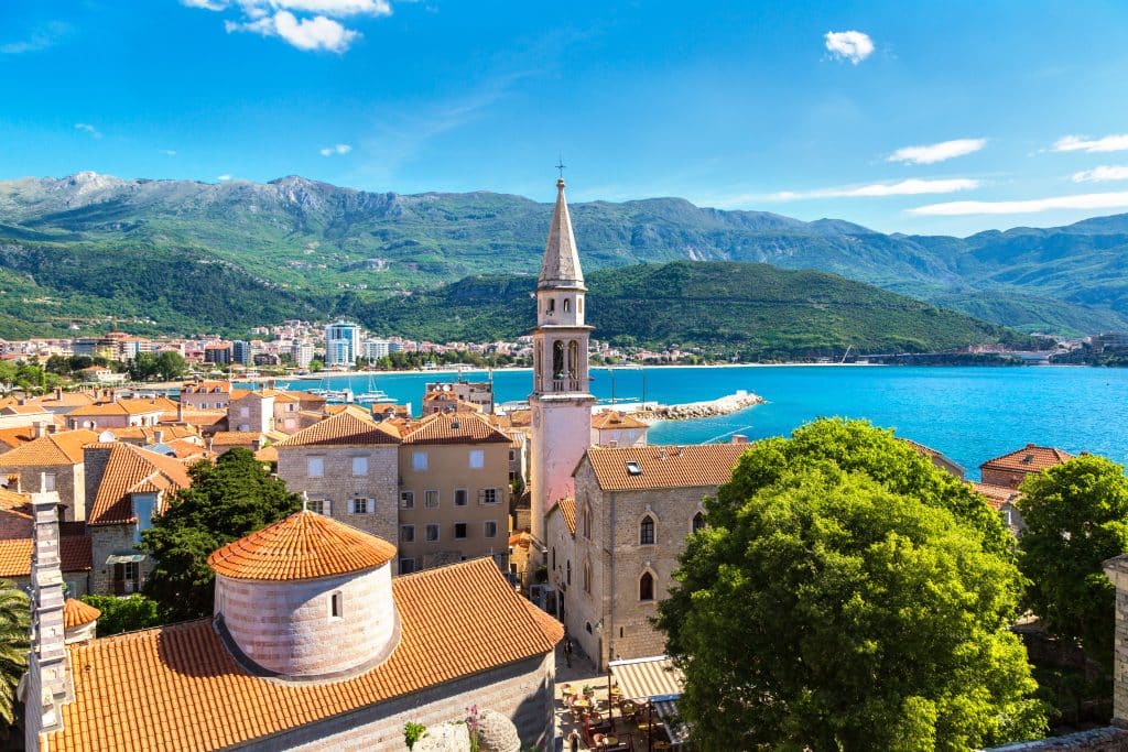 A view of the orange roofs of Budva's old town, a church tower the tallest of all of them, with the sea in the background.