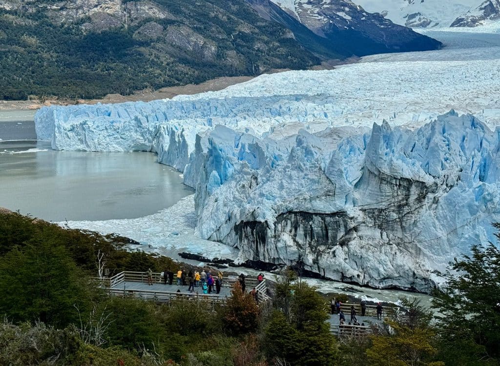 An enormous blue and white glacier with crowds standing on platforms watching it.