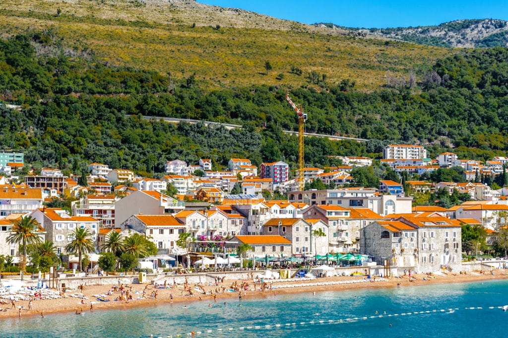 A small beach full of people in front of a town with stone buildings topped with orange roofs.