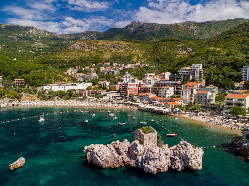 A long pebbly beach in the middle of a resort town in Montenegro surrounded by tall mountains.