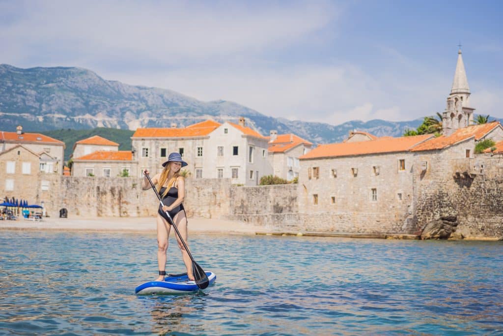 A woman in a bikini paragliding in front of the stone buildings of Budva's old town.