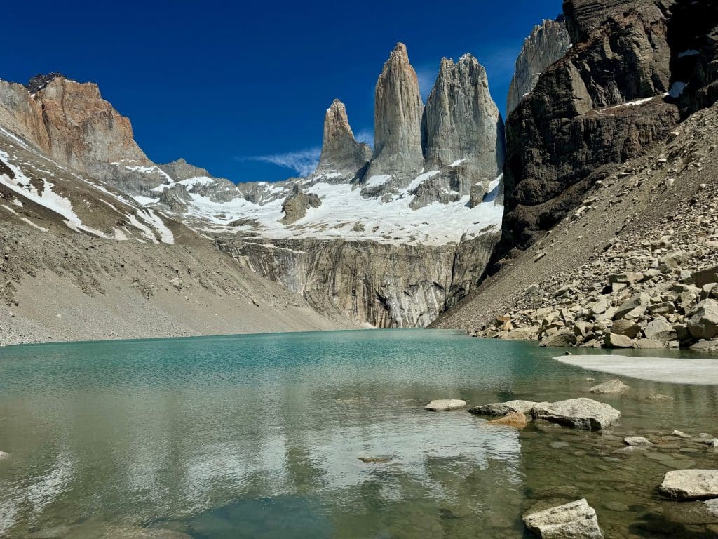 A pristine, calm green-blue lake surrounded by Rocky Mountains, including three sharp rocky spires in the distance.