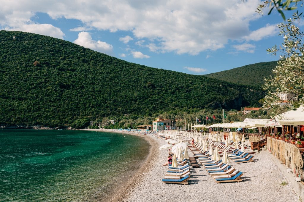 Rows of beach chairs on a white pebble beach facing a calm, dark green, clear sea.