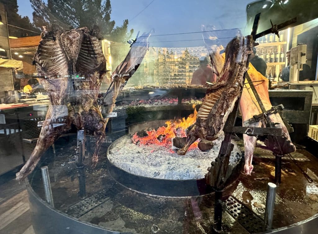 The view through a window of several large pieces of lamb roasting on a fire.