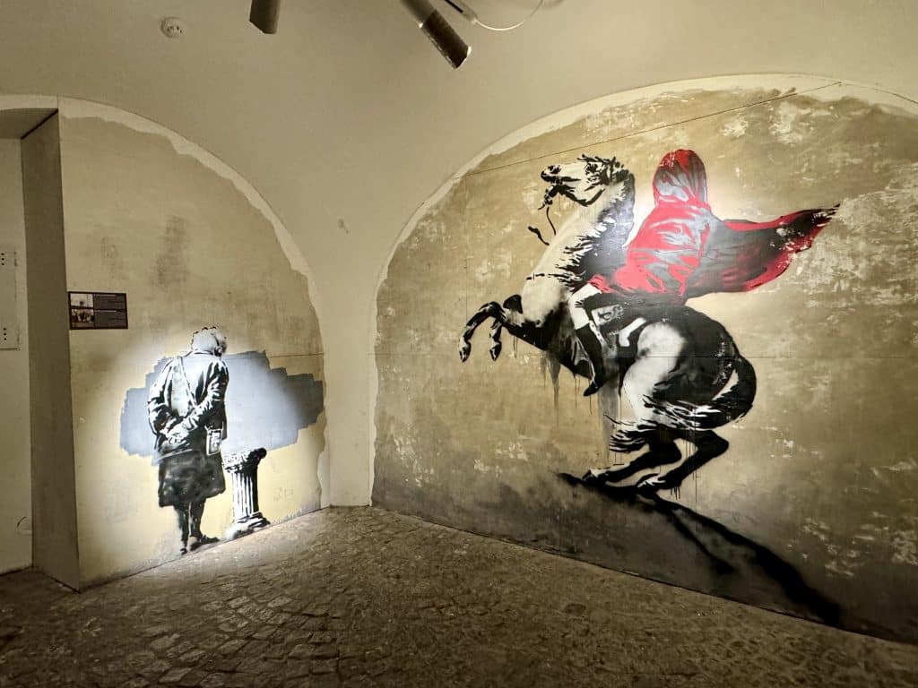 Cement walls with Banksy's art painted on them, including a figure riding a horse with his cloak obscuring his face.