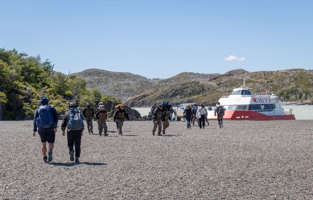 Several people walking up a gray sand beach where a boat is waiting for them to board.