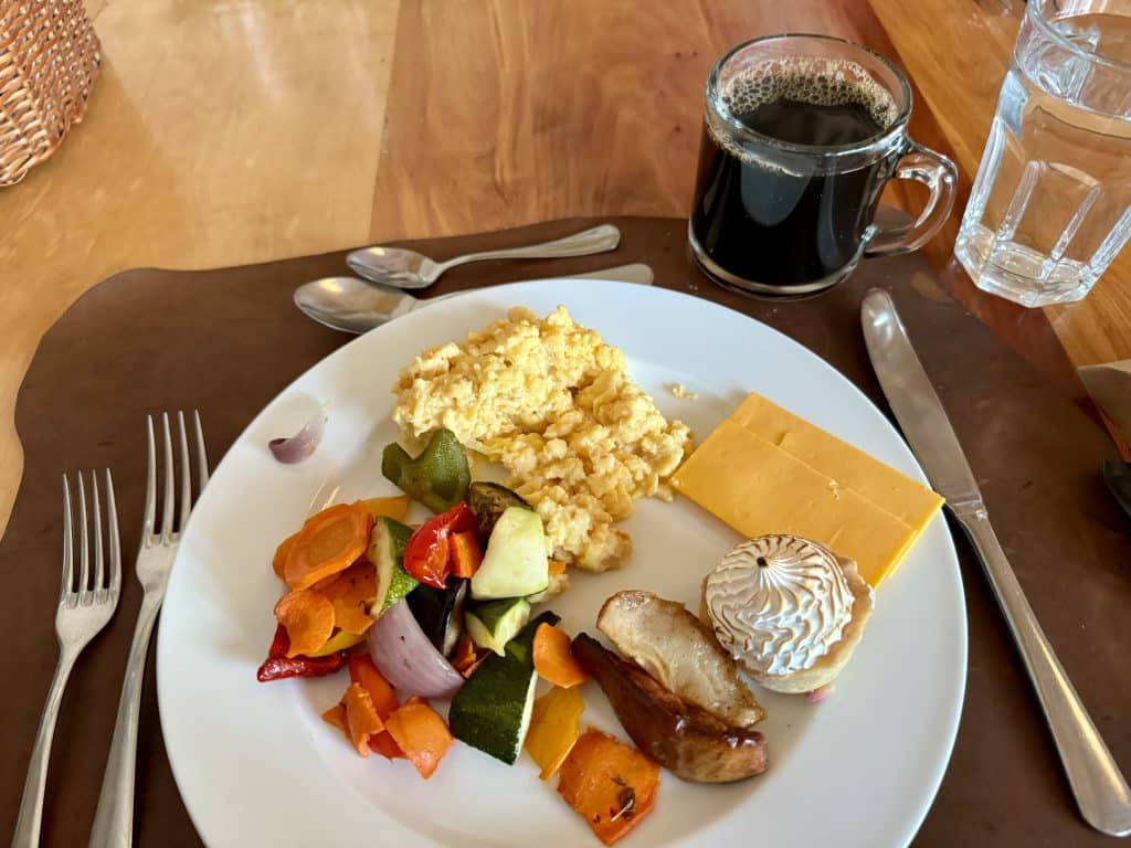 A breakfast plate with scrambled eggs, vegetables, a tiny lemon meringue pastry, and some cheese, next to a cup of black coffee.