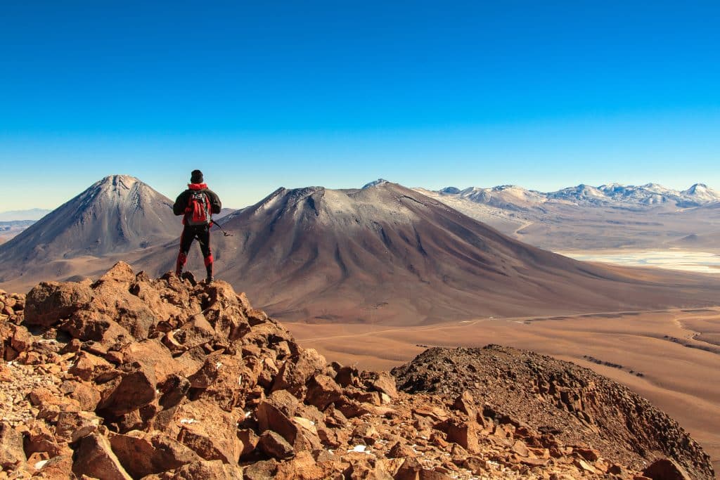 A man climbing a mountain in the Atacama Desert, with views of mountains underneath a bright blue sky around him.