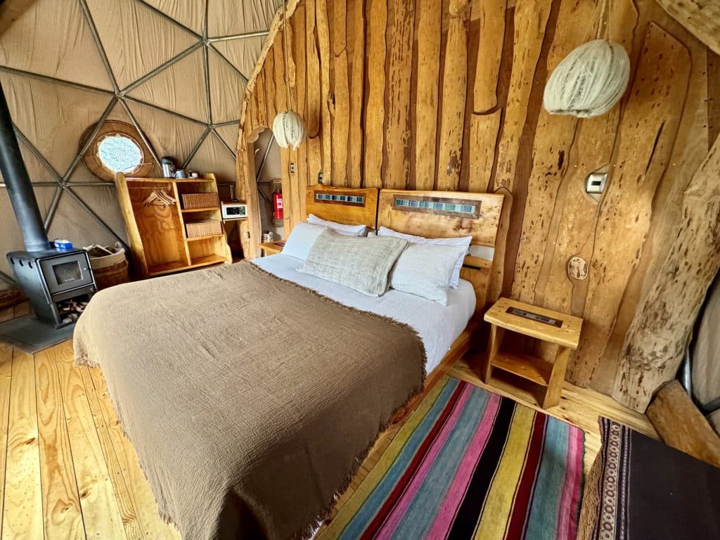 A dome hotel room with a big bed covered with blankets and surrounded by colorful rugs. Behind it is a wooden headboard made out of tree trunks.