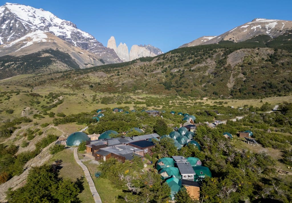 A drone view of Ecocamp Patagonia -- a cluster of small green domes in the middle of a hilly, grassy area, with a snowy mountain and the three jagged gray rock towers in the distance behind it.
