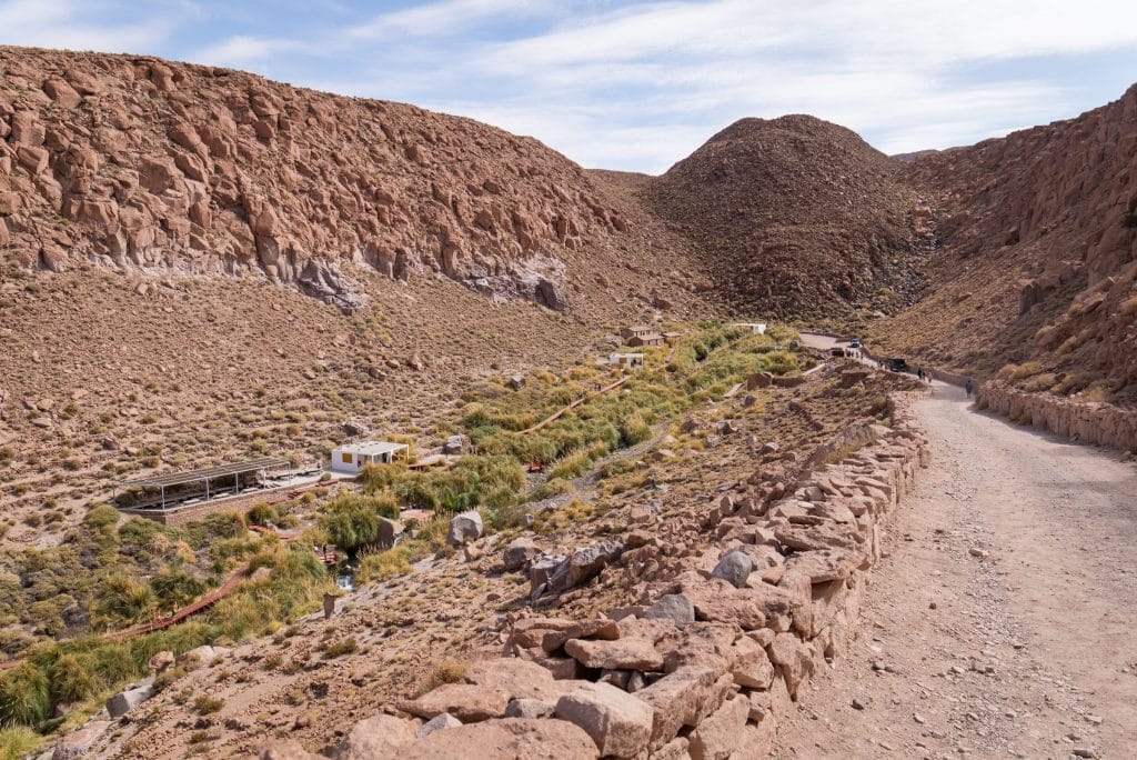 A dirt road leading downward into a rocky canyon, with a rushing river and a surprising amount of vegetation surrounding it.