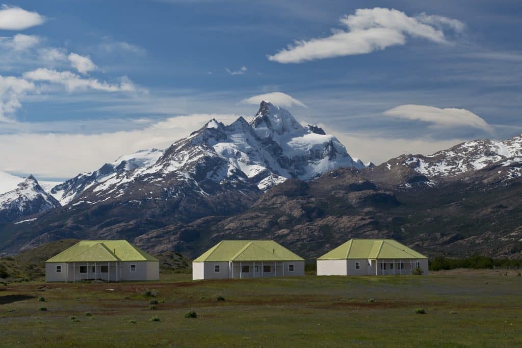 Three low buildings with green roofs in the Patagonian countryside, a pointy mountain rising behind them.