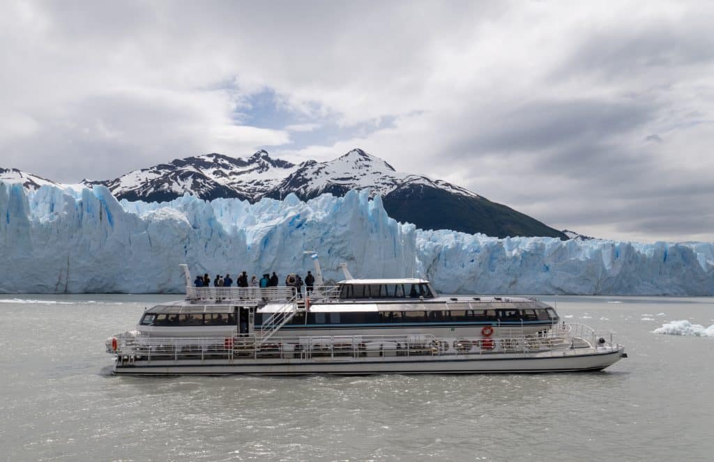 A large boat cruising in front of a massive blue and white glacier, snow-capped mountains behind it.