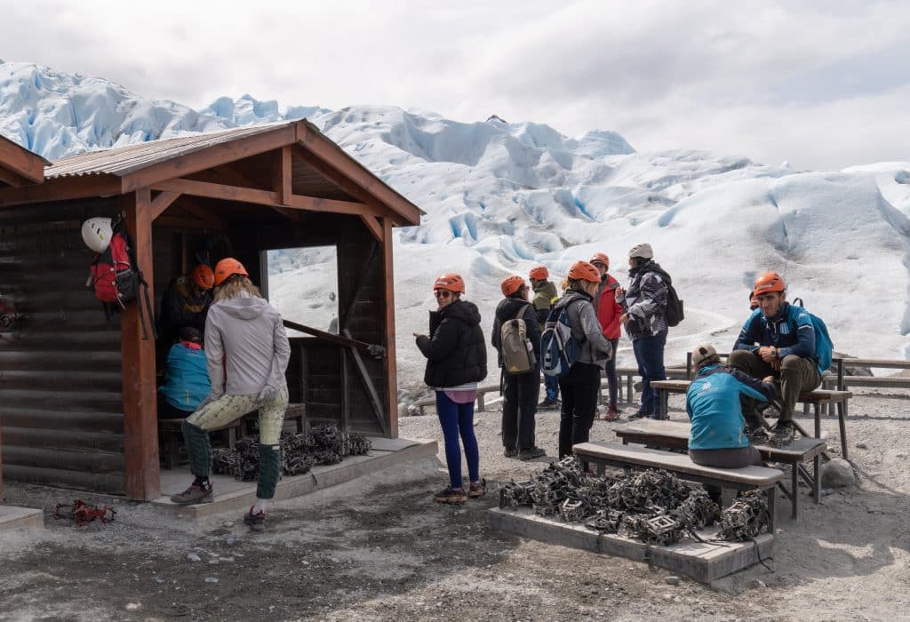 A group of hikers clustered around a small cabin where guides are tying crampons on their boots.