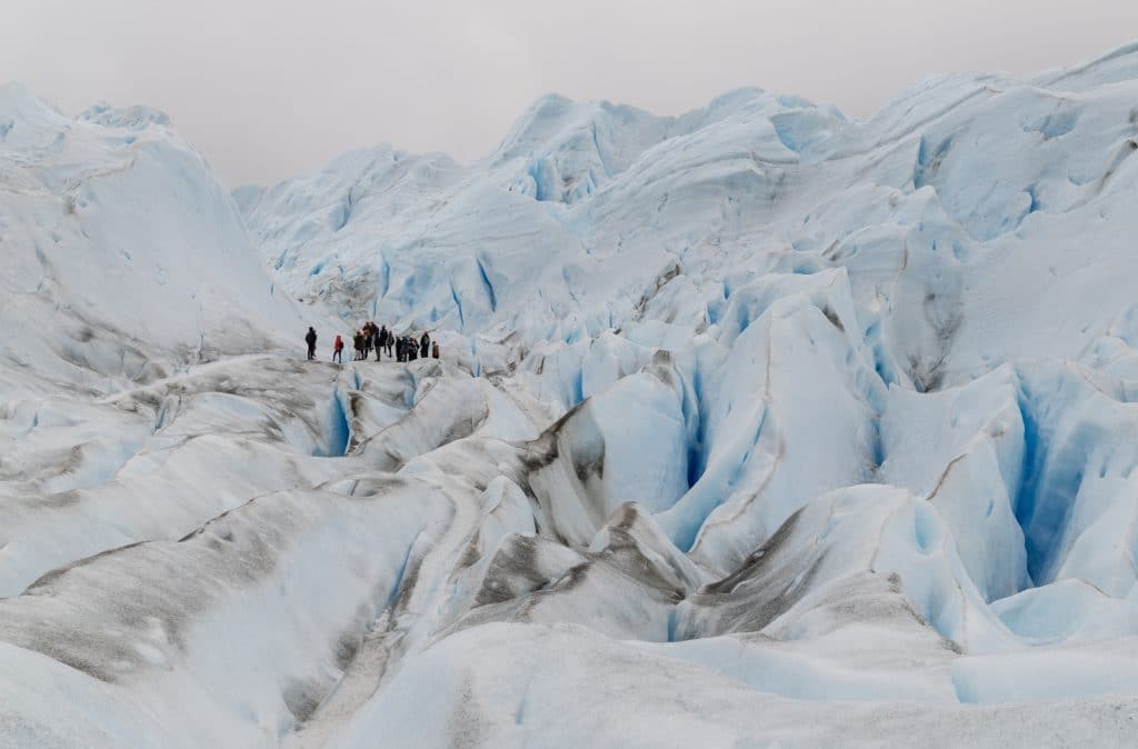 A group of tiny people clustered in a small corner of a massive blue and white glacier, showing you how enormous the glacier is.