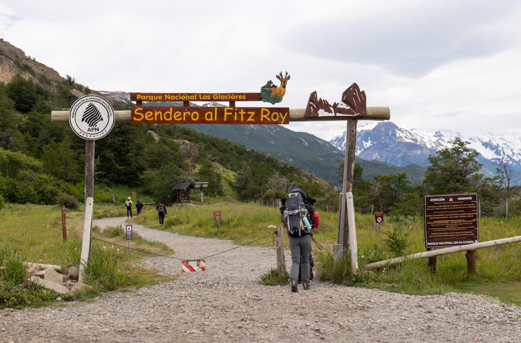A hiker with a backpack walking underneath a wooden gate reading "Sendero al Fitz Roy."