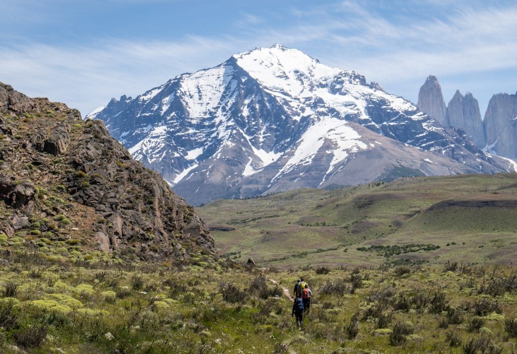 A gorgeous hiking view with several people hiking through a green valley, a huge gray snow-capped mountain rising in the distance.